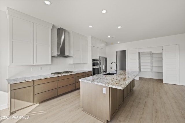 kitchen featuring a kitchen island with sink, sink, wall chimney exhaust hood, light wood-type flooring, and white cabinetry