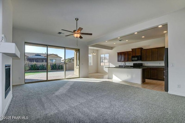 kitchen with light carpet, ceiling fan with notable chandelier, dark brown cabinetry, black appliances, and a center island with sink