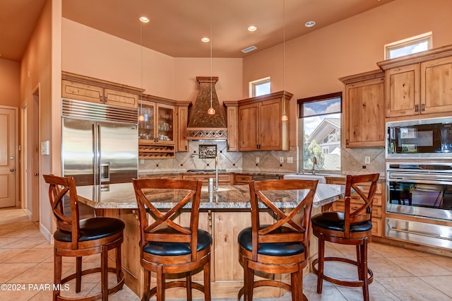 kitchen featuring light stone counters, built in appliances, a breakfast bar area, a kitchen island with sink, and custom exhaust hood