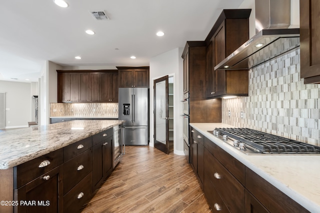 kitchen with appliances with stainless steel finishes, light wood-type flooring, light stone counters, dark brown cabinets, and wall chimney range hood