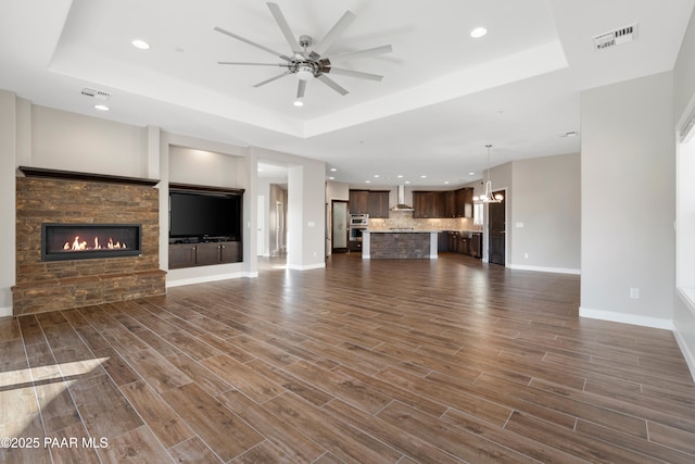 unfurnished living room with a raised ceiling, a stone fireplace, and ceiling fan with notable chandelier