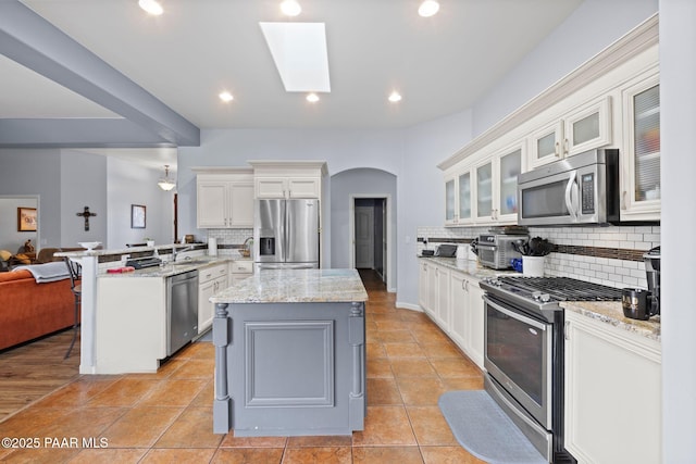 kitchen featuring glass insert cabinets, light stone counters, appliances with stainless steel finishes, a peninsula, and arched walkways