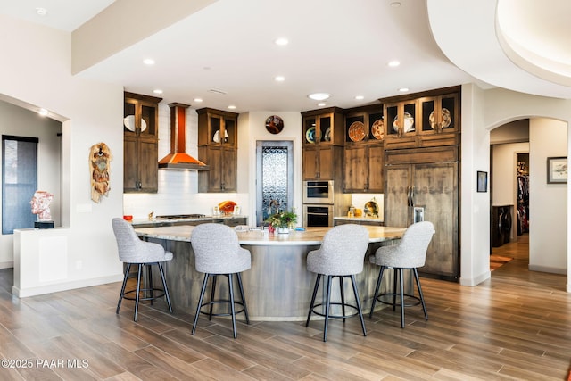 kitchen with a kitchen bar, appliances with stainless steel finishes, wall chimney exhaust hood, a spacious island, and hardwood / wood-style flooring