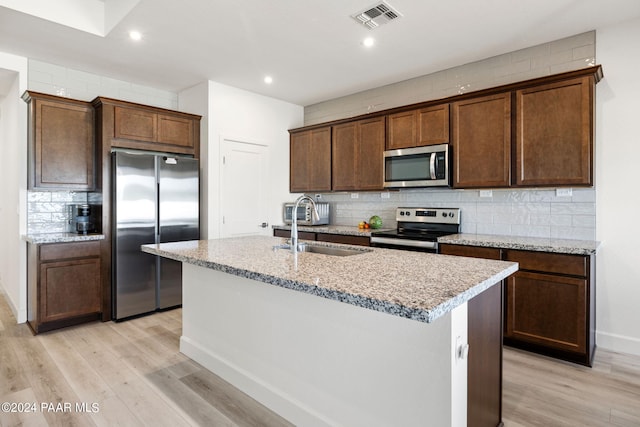 kitchen with light stone countertops, light wood-type flooring, stainless steel appliances, sink, and a center island with sink