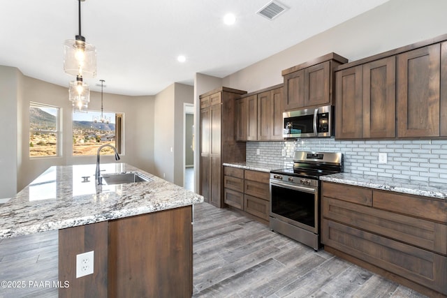 kitchen with light stone countertops, appliances with stainless steel finishes, a kitchen island with sink, sink, and decorative light fixtures