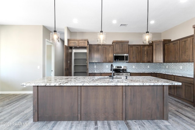 kitchen featuring light stone counters, dark brown cabinetry, stainless steel appliances, a center island with sink, and light hardwood / wood-style floors