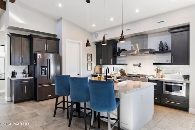 kitchen with appliances with stainless steel finishes, hanging light fixtures, wall chimney exhaust hood, and an island with sink