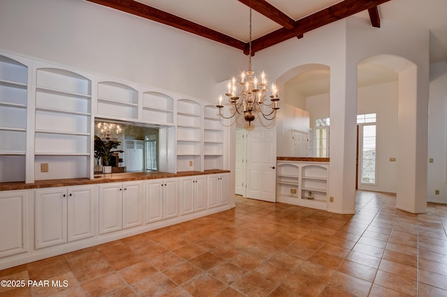 unfurnished dining area featuring built in features, beam ceiling, light tile patterned floors, a high ceiling, and an inviting chandelier