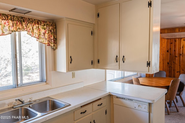 kitchen featuring a sink, a peninsula, wood walls, white dishwasher, and light countertops