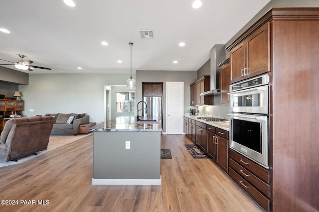 kitchen featuring appliances with stainless steel finishes, wall chimney range hood, pendant lighting, light hardwood / wood-style flooring, and an island with sink