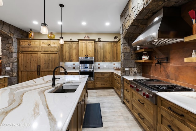 kitchen featuring light stone counters, wall chimney range hood, decorative backsplash, appliances with stainless steel finishes, and sink