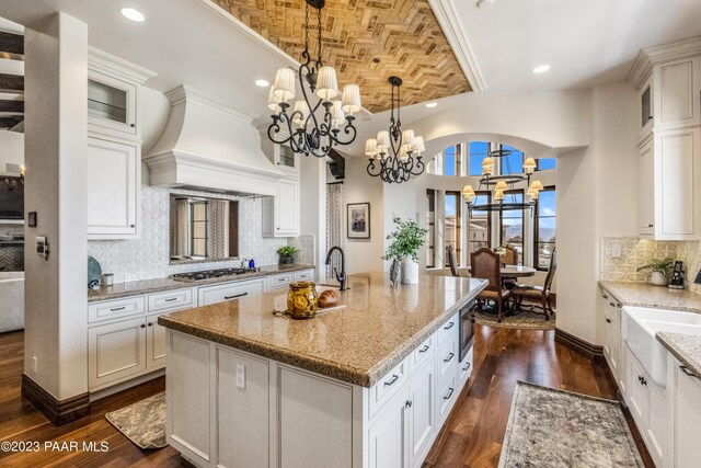 kitchen with dark wood-type flooring, an inviting chandelier, crown molding, an island with sink, and custom exhaust hood