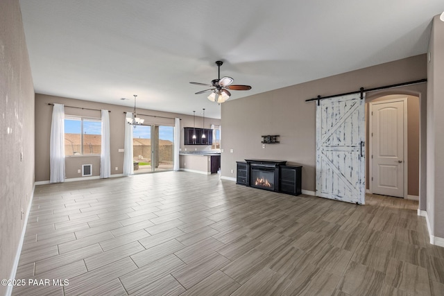 unfurnished living room featuring ceiling fan with notable chandelier, a glass covered fireplace, a barn door, light wood finished floors, and baseboards