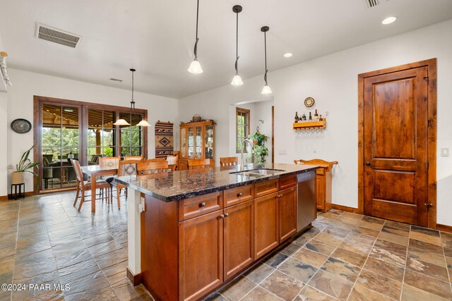 kitchen with dishwasher, a center island with sink, sink, hanging light fixtures, and dark stone countertops