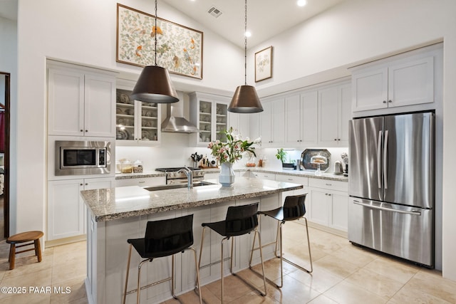 kitchen with stainless steel appliances, a sink, wall chimney range hood, light stone countertops, and a center island with sink