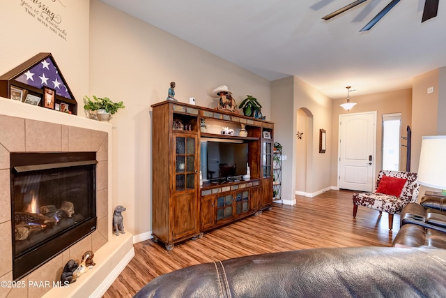 living room featuring arched walkways, a tiled fireplace, a ceiling fan, wood finished floors, and baseboards