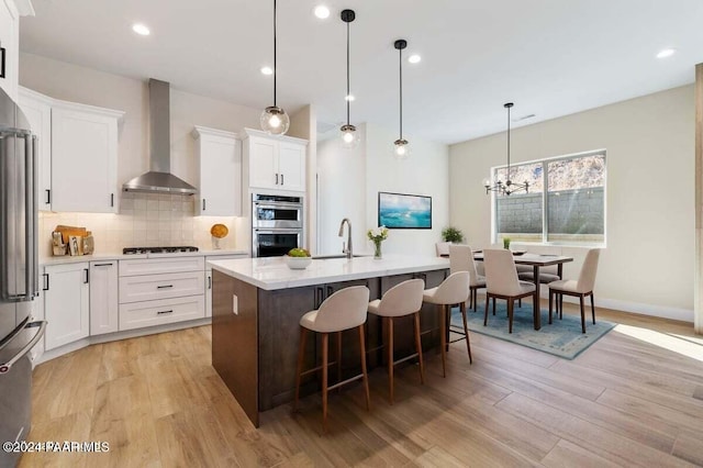 kitchen featuring an island with sink, decorative light fixtures, wall chimney range hood, and light wood-type flooring