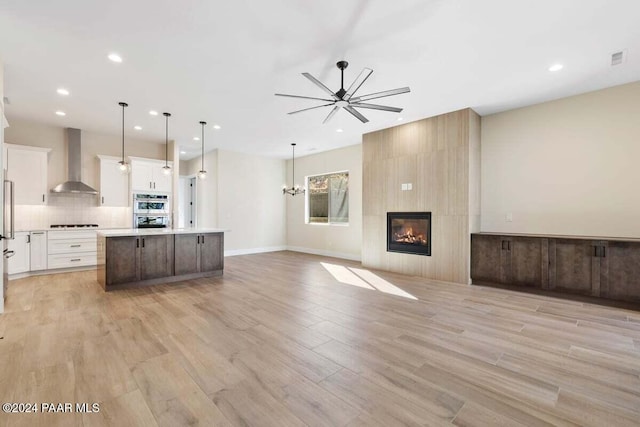 kitchen featuring white cabinets, a center island with sink, hanging light fixtures, wall chimney exhaust hood, and light wood-type flooring