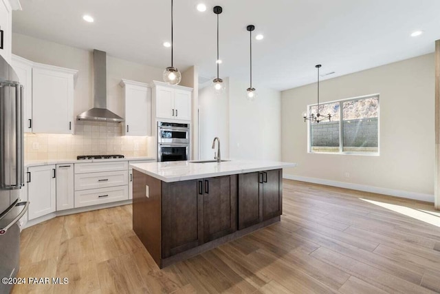 kitchen with pendant lighting, wall chimney range hood, an island with sink, and light hardwood / wood-style flooring