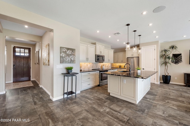 kitchen featuring a kitchen island with sink, dark stone counters, sink, appliances with stainless steel finishes, and decorative light fixtures