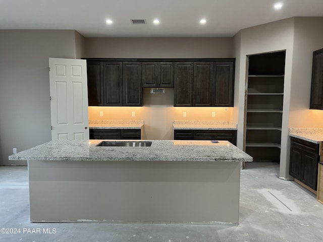 kitchen featuring dark brown cabinets, light stone counters, and a kitchen island with sink