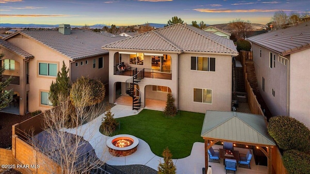 back house at dusk featuring a lawn, a gazebo, a patio area, an outdoor living space with a fire pit, and a balcony