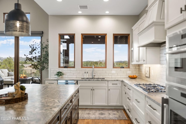 kitchen with stainless steel gas cooktop, custom exhaust hood, a sink, white cabinetry, and a wealth of natural light