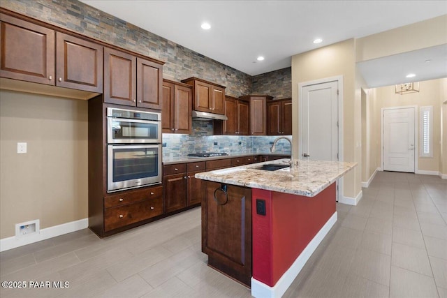 kitchen featuring sink, a kitchen island with sink, stainless steel appliances, light stone counters, and decorative backsplash