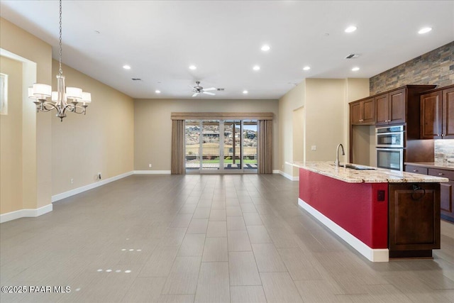 kitchen with sink, a center island with sink, double oven, pendant lighting, and light stone countertops