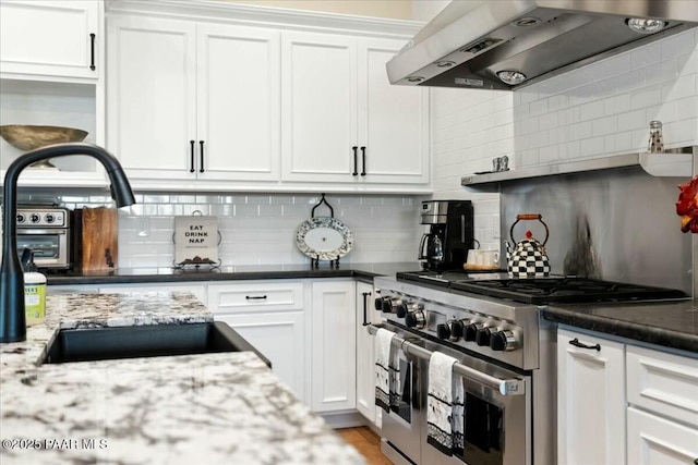 kitchen with wall chimney range hood, dark stone countertops, high end stove, white cabinets, and decorative backsplash