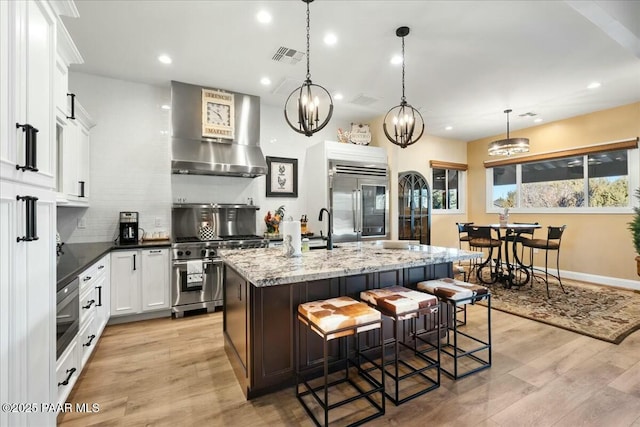 kitchen featuring decorative light fixtures, high quality appliances, white cabinets, wall chimney range hood, and a center island with sink