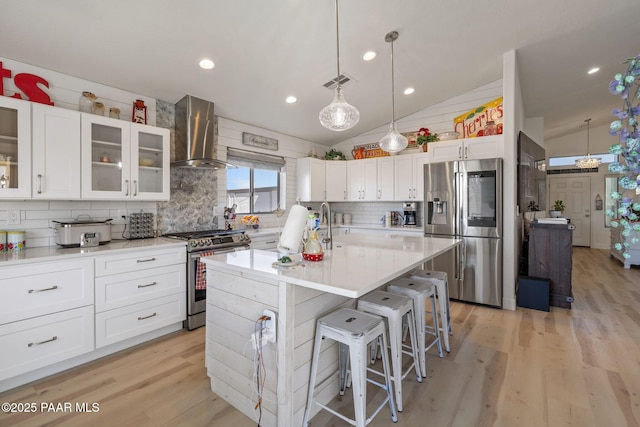 kitchen with light wood-style flooring, stainless steel appliances, vaulted ceiling, white cabinetry, and wall chimney range hood