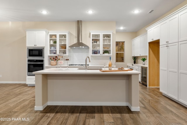 kitchen with stainless steel oven, an island with sink, white cabinets, built in microwave, and wall chimney exhaust hood