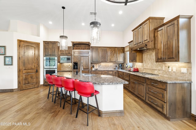 kitchen featuring stone counters, an island with sink, built in appliances, and decorative light fixtures