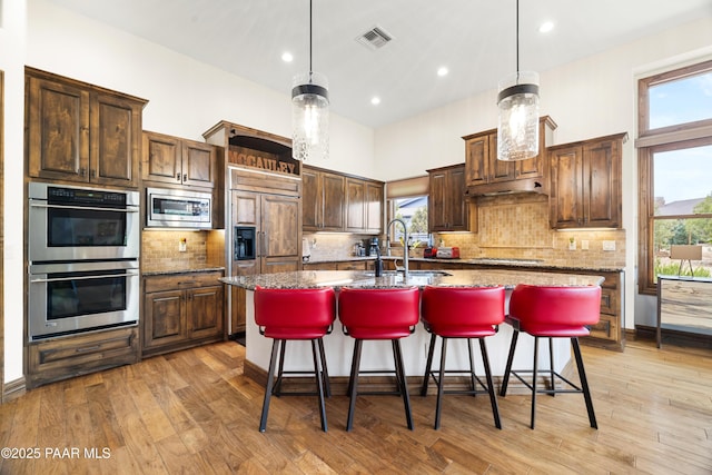 kitchen featuring a center island with sink, pendant lighting, built in appliances, and dark stone counters