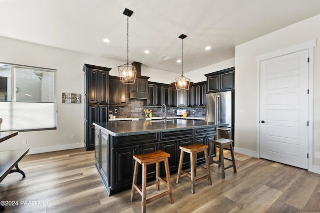 kitchen featuring light wood finished floors, dark countertops, high quality fridge, a kitchen island with sink, and backsplash