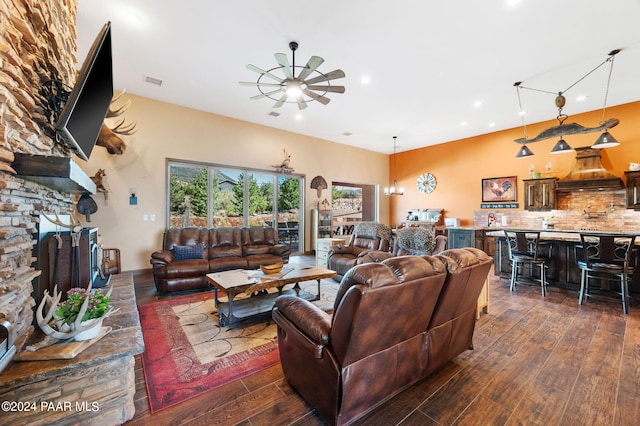 living room with ceiling fan with notable chandelier, a stone fireplace, and dark wood-type flooring