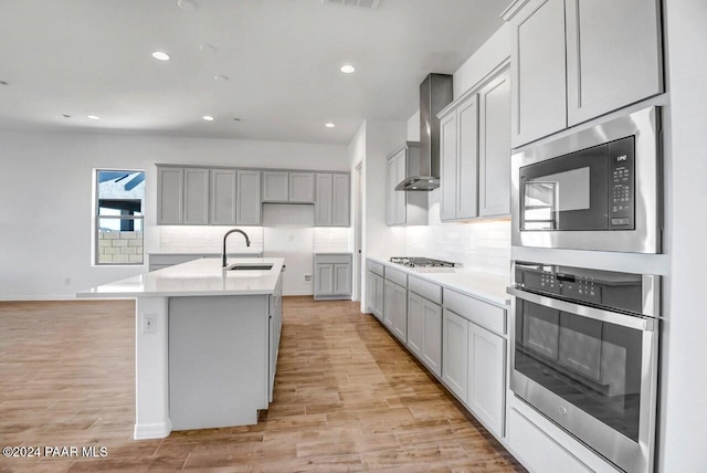 kitchen with appliances with stainless steel finishes, light wood-type flooring, wall chimney exhaust hood, sink, and a center island with sink