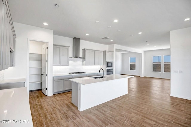 kitchen with a center island with sink, light wood-type flooring, wall chimney range hood, and appliances with stainless steel finishes