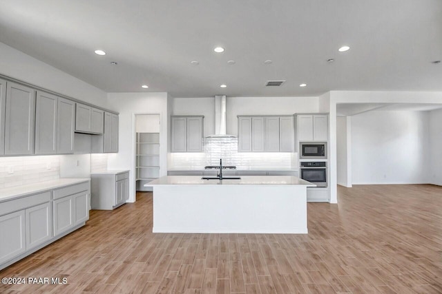 kitchen featuring a kitchen island with sink, oven, wall chimney exhaust hood, light wood-type flooring, and black microwave