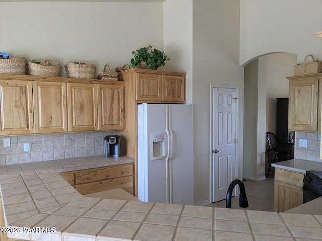 kitchen featuring tile patterned flooring, white fridge with ice dispenser, tile counters, light brown cabinetry, and tasteful backsplash