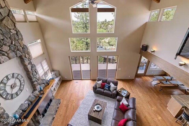 living room featuring beamed ceiling, ceiling fan, light hardwood / wood-style flooring, a high ceiling, and a stone fireplace