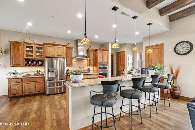 kitchen with light stone countertops, stainless steel appliances, wall chimney range hood, wood-type flooring, and hanging light fixtures