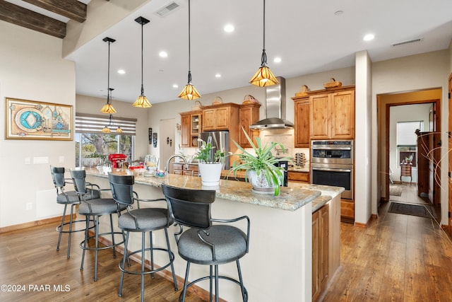 kitchen featuring a large island, wall chimney exhaust hood, light hardwood / wood-style flooring, pendant lighting, and a kitchen bar