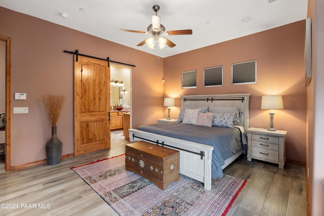 bedroom with ensuite bathroom, a barn door, light wood-type flooring, and ceiling fan