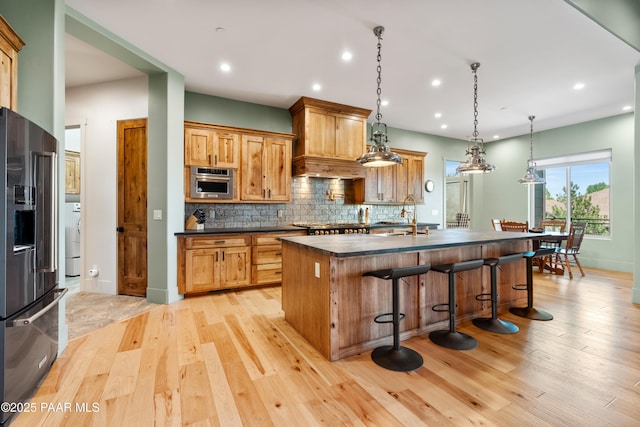 kitchen featuring a kitchen island with sink, stainless steel appliances, dark countertops, and light wood-type flooring