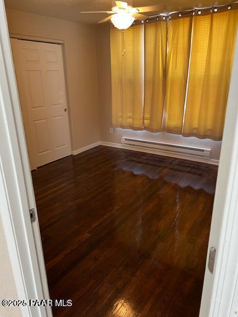 empty room featuring dark wood-style floors, baseboard heating, baseboards, and a ceiling fan