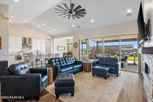 living room with light wood-type flooring, ceiling fan, sink, high vaulted ceiling, and a stone fireplace