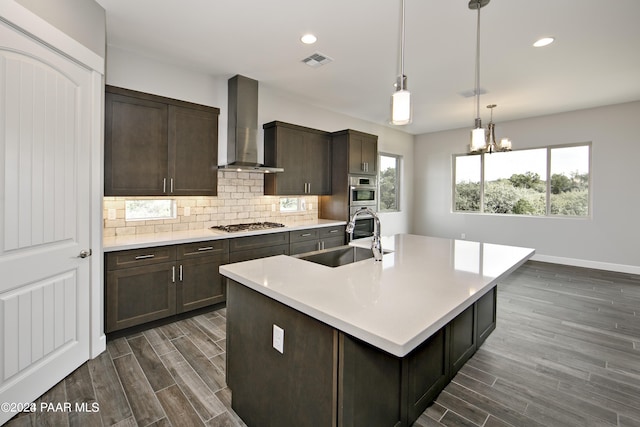 kitchen with sink, backsplash, dark brown cabinetry, a center island with sink, and wall chimney exhaust hood