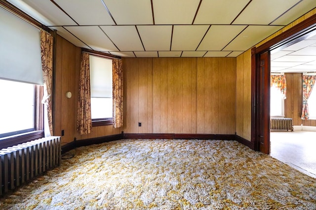 carpeted empty room with wood walls, radiator heating unit, and a drop ceiling